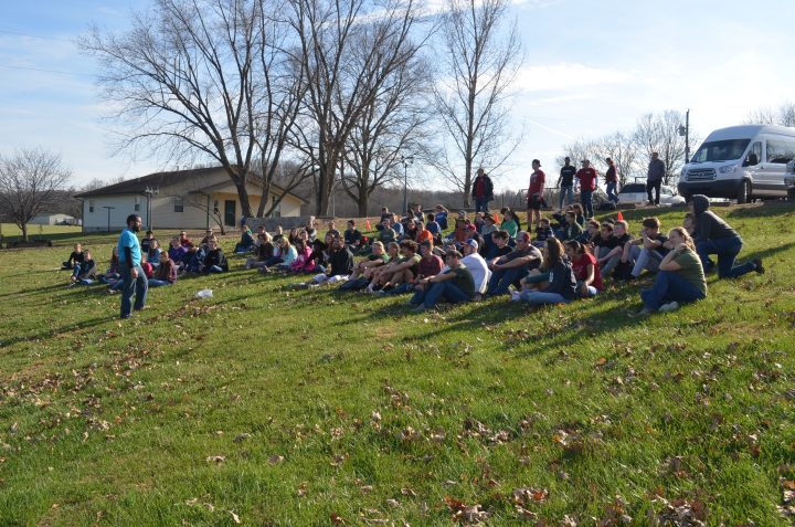 Image of a group of campers sitting on a grassy hill and listening to the director speak
